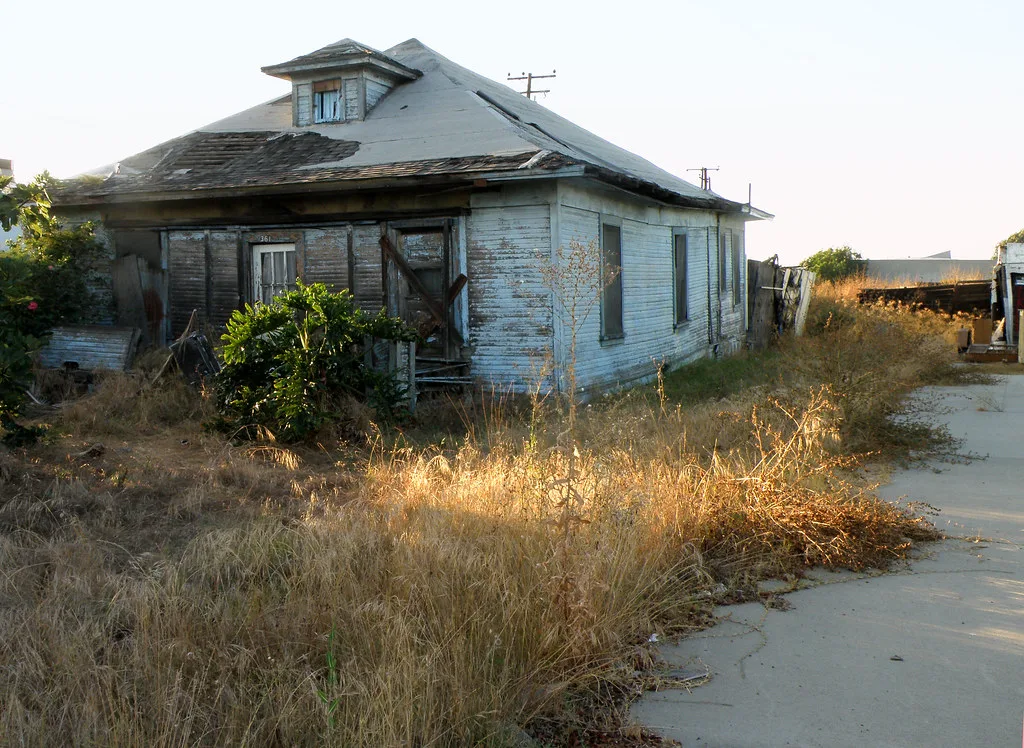 Abandoned pale-blue wooden house with a damaged roof amid dry grass.