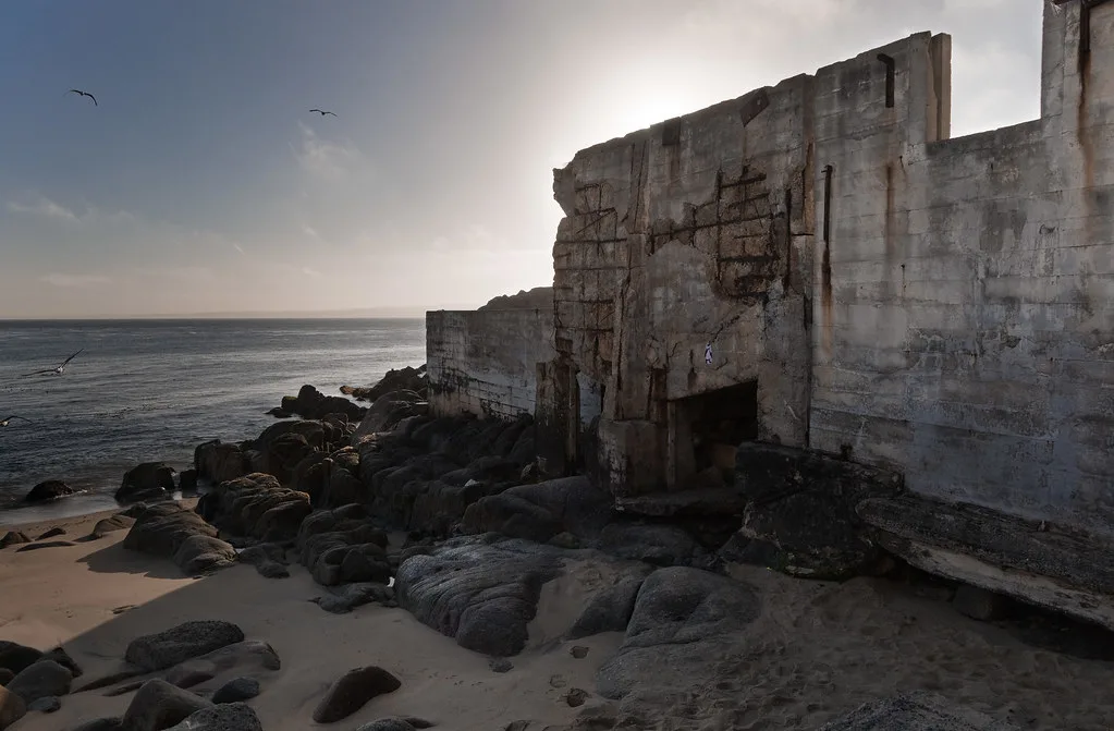 Ruines de murs de béton au bord d’une plage rocheuse.