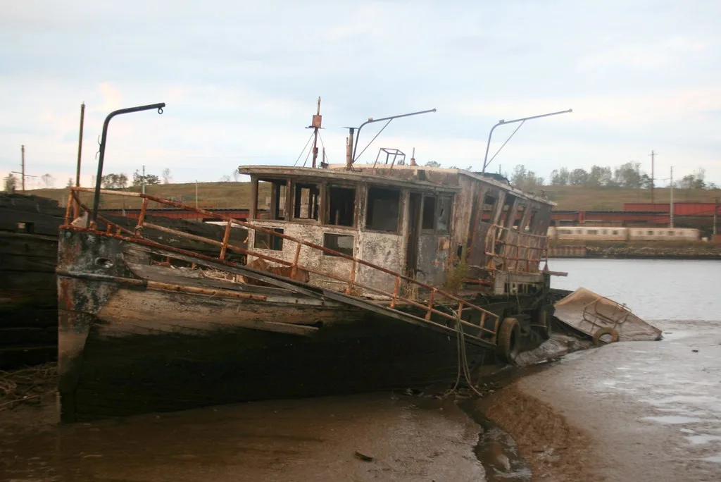 Leaning wooden boat wreck in the mud, with a gutted cabin and rusty railings.