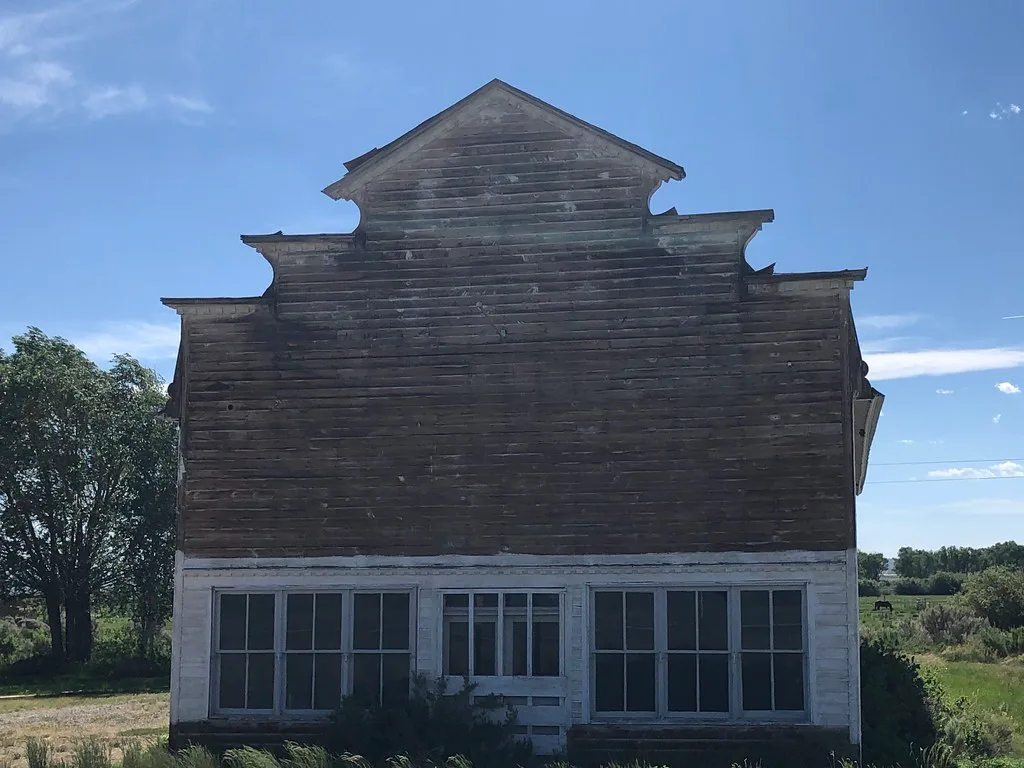 Former wooden shop with a stepped false front, weathered door and vegetation across the facade.
