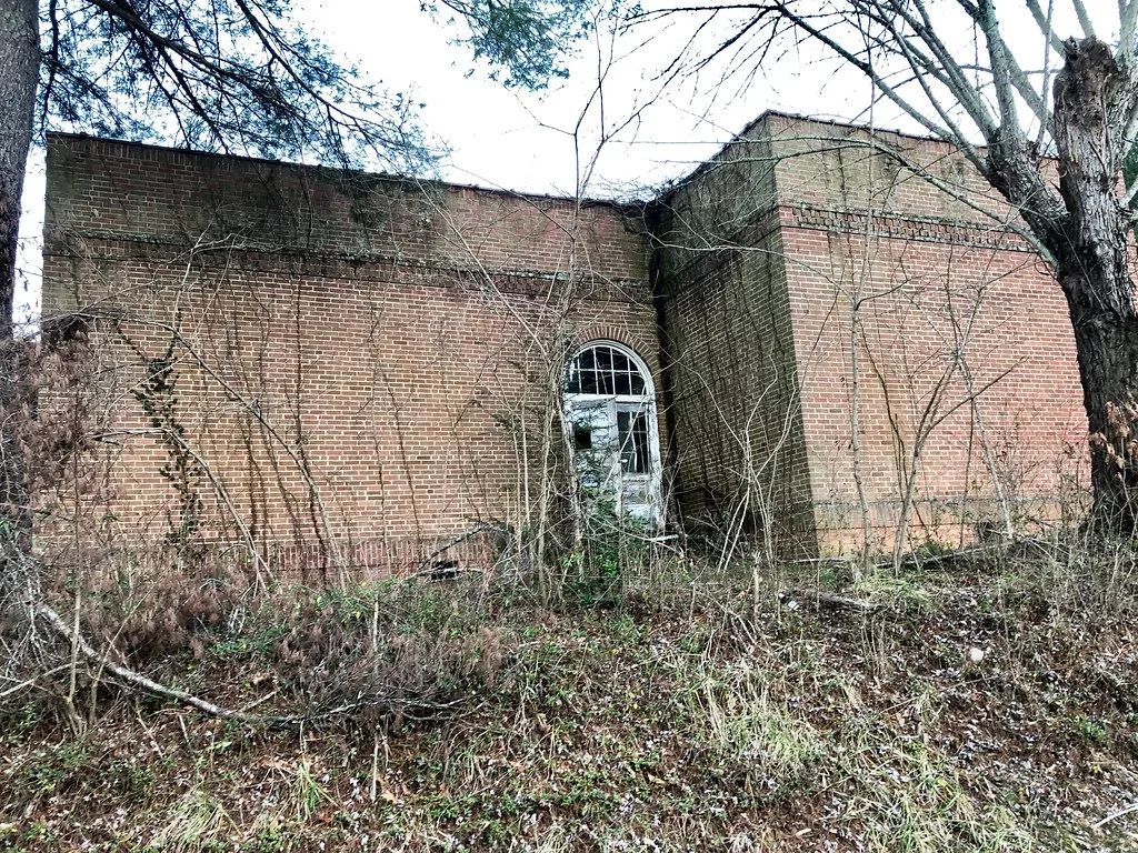 Abandoned brick building with a broken glazed arched door behind trees and brush.