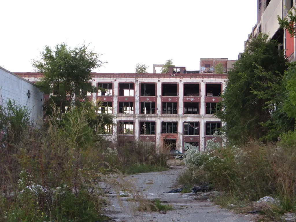 Factory courtyard overgrown with shrubs facing a red brick building with glassless windows.