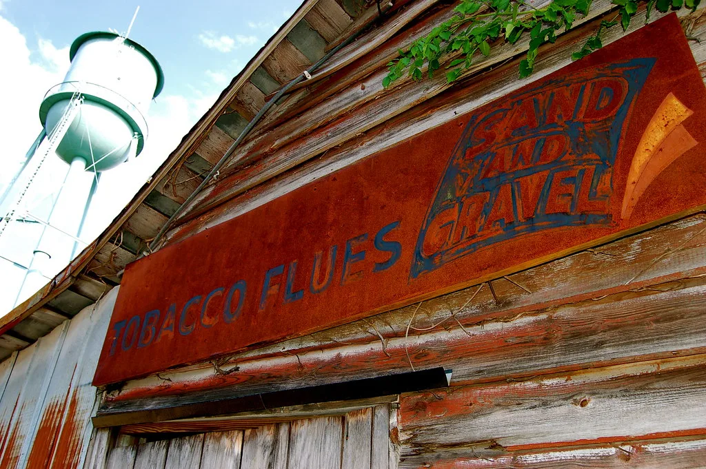 Old rusty sign on weathered wooden cladding, with a water tower behind it.