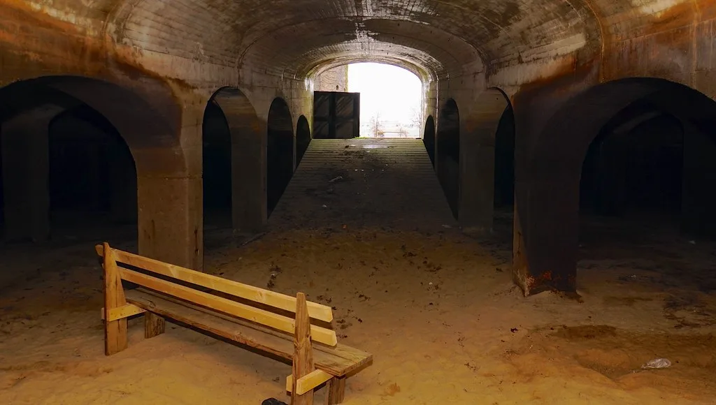Galerie voûtée aux ouvertures latérales sombres et au sol couvert de sable.