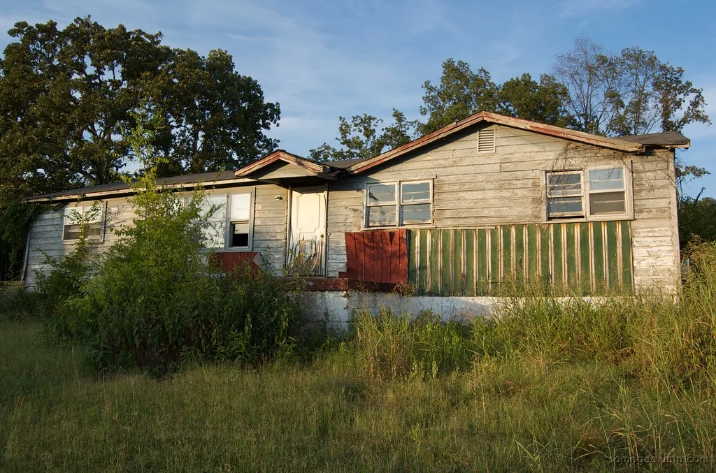 Abandoned light-clad house with a red and green porch surrounded by tall grass.