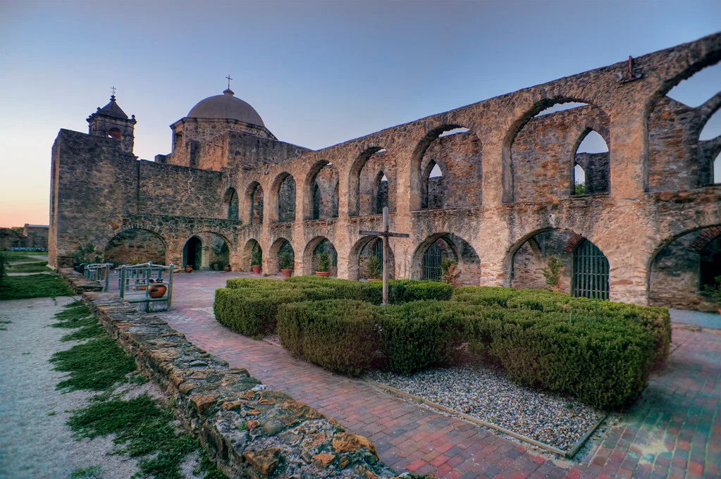 Roofless two-storey stone arcade ruins around a planted courtyard beside a domed church.