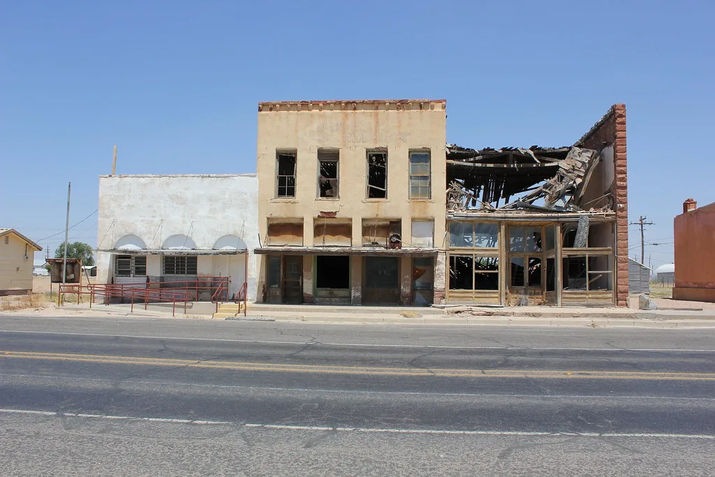 Abandoned shops with shattered storefronts and collapsed roof framing beside a road.