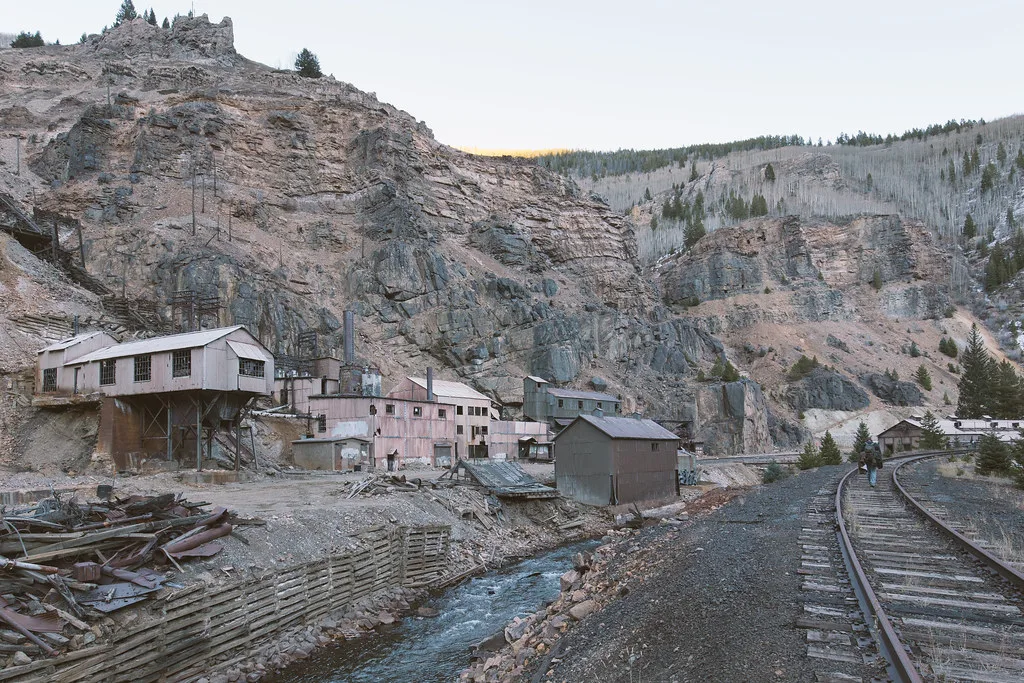 Industrial buildings with broken windows below a cliff beside a stream and railway.